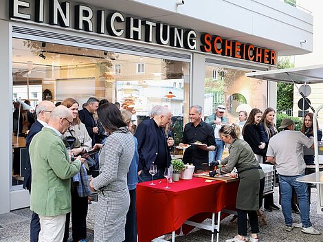 People socializing at an outdoor event in front of Einrichtung Scheicher store