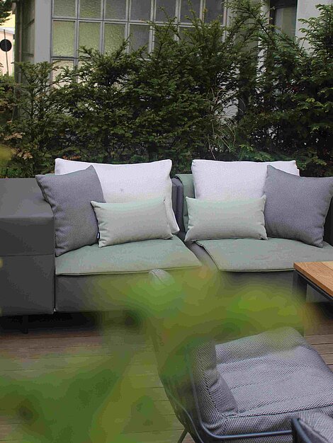 Moderne Gartensofas mit grauen Kissen auf einer Holzterrasse vor grünen Hecken
