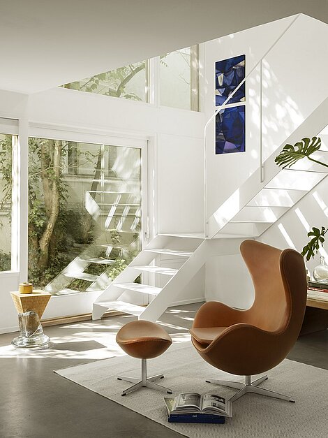 Modernes Wohnzimmer mit braunem Egg Chair und weißer Treppe in heller, minimalistischer Einrichtung