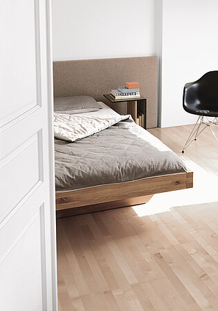 Modernes Schlafzimmer mit Holzbett, grauer Bettwäsche und Designstuhl auf hellem Parkettboden