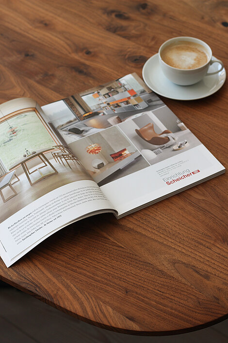 Geöffnete Wohnzeitschrift mit Einrichtungsideen neben einer Tasse Cappuccino auf Holztisch