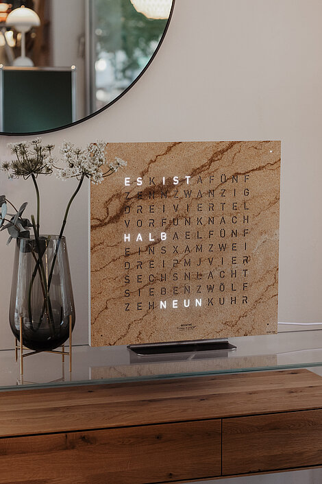 Moderne Wortuhr mit Steinoptik auf Holzkommode neben Vase mit weißen Blumen