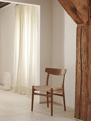 Holzstuhl mit geflochtener Sitzfläche in minimalistischem Wohnzimmer mit Holzbalken und Vorhang