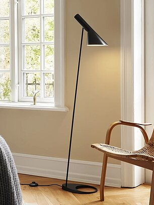 Schwarze Stehlampe neben geflochtenem Holzstuhl in hellem Wohnzimmer mit großen Fenstern