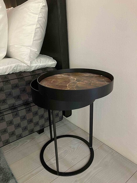 Runder Beistelltisch mit dunkler Marmorplatte und schwarzem Metallgestell neben einem Bett