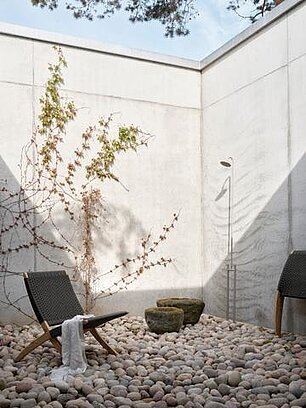Minimalistische Außendusche mit Kiesboden, zwei Liegestühlen und Kletterpflanze an Betonwand