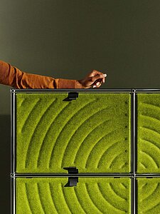 Modulares Regal mit grüner Filzfront in Kreismuster neben Arm in braunem Pullover