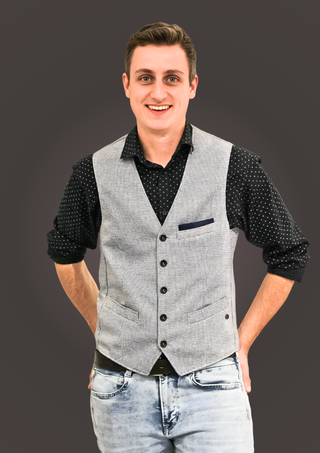 Lächelnder junger Mann in Jeans, schwarzem Hemd mit Punkten und grauer Weste vor dunklem Hintergrund