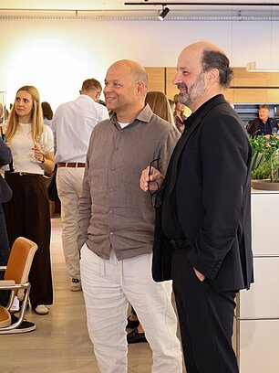 Two men in casual attire at a social event, surrounded by people with drinks