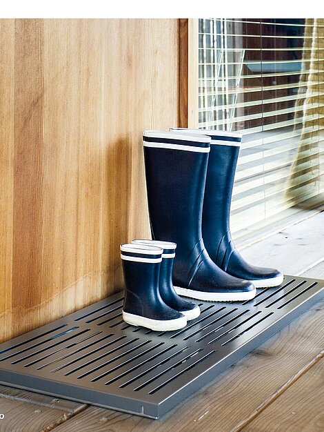 Two pairs of black rubber boots on a metal mat by a wooden wall
