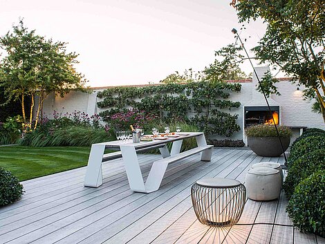 Moderne Gartenterrasse mit weißer Sitzbank, Esstisch und Außenkamin bei Abendlicht