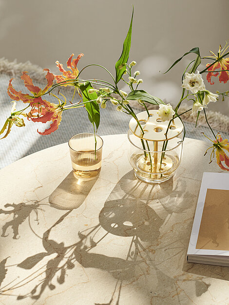 Glasvase mit exotischen Blumen auf Marmortisch im Sonnenlicht neben Glas und Notizbuch