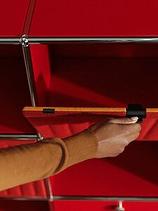 Hand öffnet rote Filztür eines modularen Regalsystems mit Metallrahmen