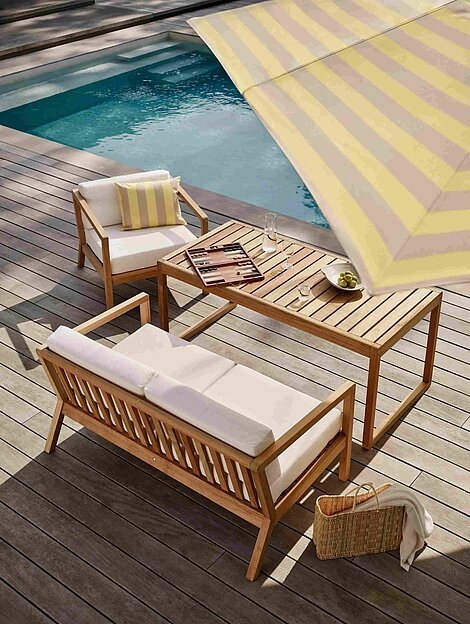 Moderne Gartenmöbel aus Holz mit Polstern auf Holzterrasse am Pool unter gestreiftem Sonnenschirm