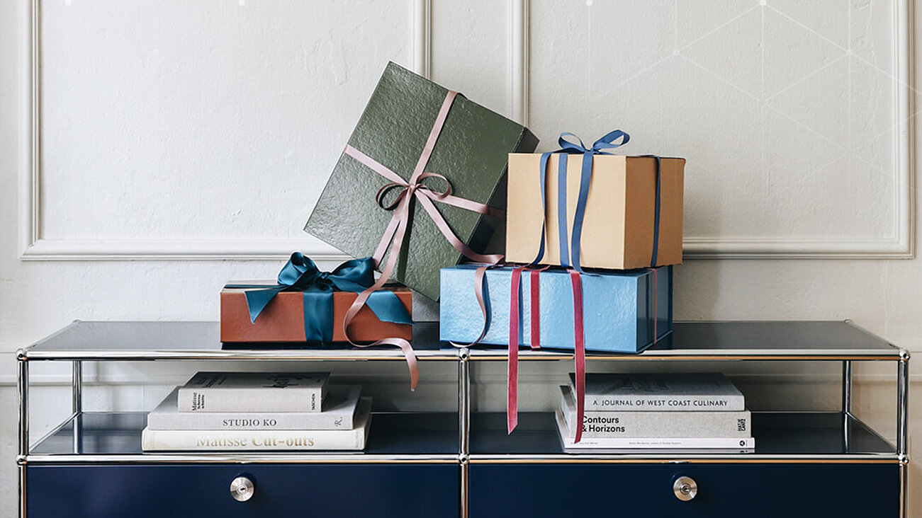 Geschenkboxen mit bunten Bändern auf dunkelblauem Metallregal neben Büchern vor weißer Wand